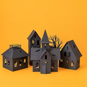 May include: A collection of black paper structures including a house with a fence, a tall tower, a central church-like building, and a house with a spider cutout. Each structure features cutouts of bats, stars, and windows with warm light.