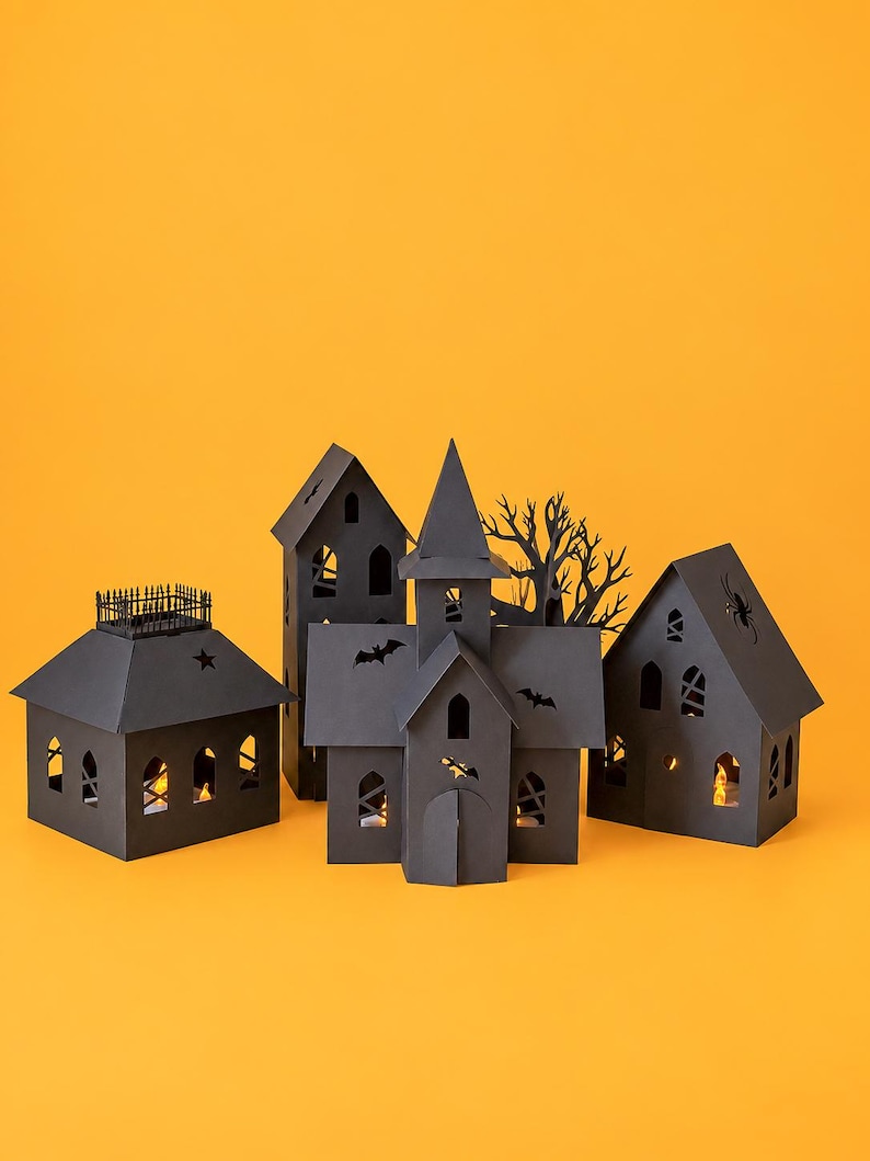 May include: A collection of black paper structures including a house with a fence, a tall tower, a central church-like building, and a house with a spider cutout. Each structure features cutouts of bats, stars, and windows with warm light.