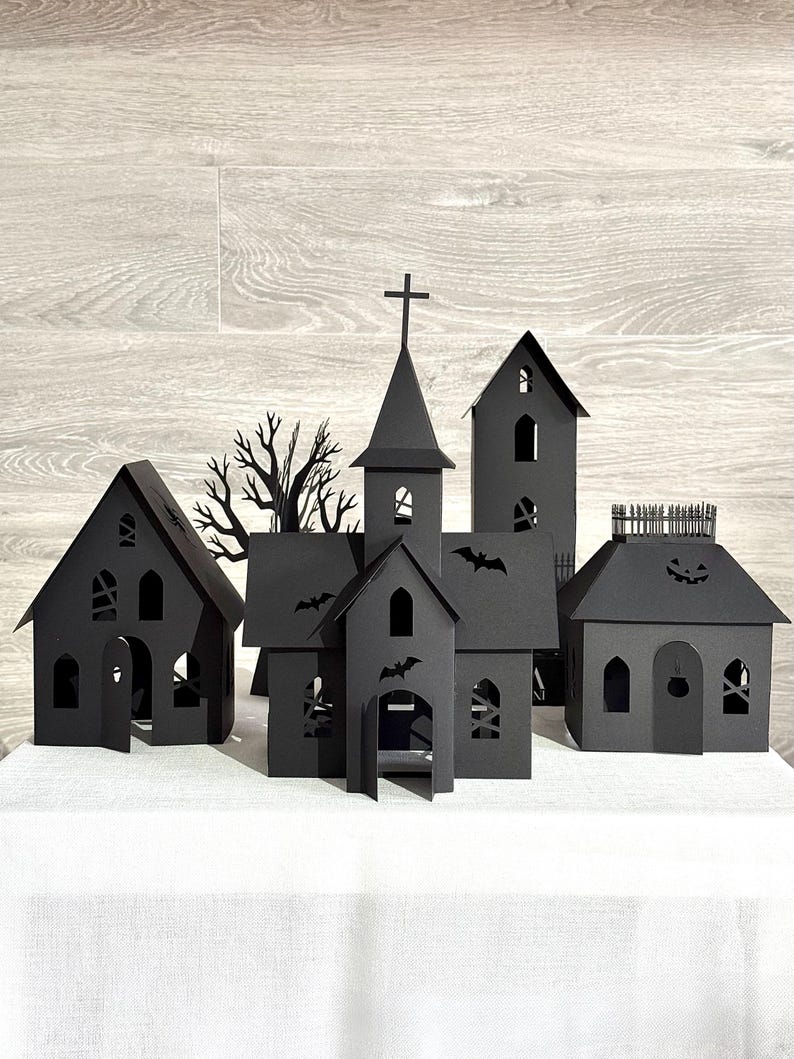 May include: A collection of black paper structures including a church with a cross, a tall tower, and houses with cut-out windows, doors, and silhouettes of bats, a bare tree, and a jack-o'-lantern face, all arranged to form a miniature village scene.