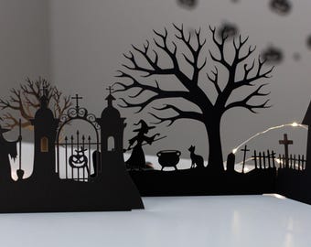 May include: Black paper Halloween decoration with silhouettes of a witch, cat, haunted house, tree, and graveyard. The scene includes a cauldron, a gate with a pumpkin, and a witch with a broom. Fairy lights are strung throughout the scene.