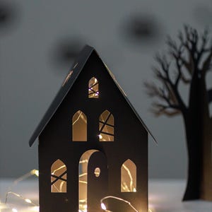 May include: A black paper house with arched windows and a partially open door, illuminated from within by warm white string lights. The structure features cut-out details on the roof and walls, creating a glowing effect through the openings.