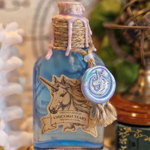 May include: A blue glass apothecary bottle labelled "Unicorn Tears" with a unicorn illustration. The bottle is sealed with a cork, twine, and pink wax. A blue and iridescent wax seal with a unicorn image hangs from the bottle.