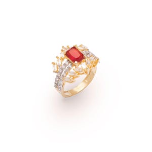 May include: A gold-toned ring featuring a large, rectangular red gemstone at the center, surrounded by smaller, clear, baguette-cut stones. The ring has a detailed, ornate design with a band that tapers towards the back. The ring is set against a white background.