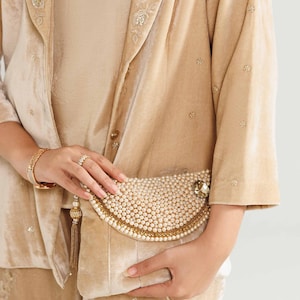 Puede incluir: Chaqueta de terciopelo beige y clutch a juego. La chaqueta tiene bordados florales en las solapas y adornos dispersos. El clutch está adornado con perlas y ribetes dorados, y una borla dorada. El conjunto es de color neutro.