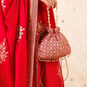 Könnte beinhalten: Ein roter, bestickter Beutel mit floralem Muster, gehalten von einer Hand. Die Tasche hat goldene Akzente und einen Perlenhenkel. Der Hintergrund zeigt roten Stoff mit goldener Stickerei.