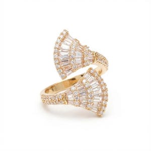 May include: A gold-toned ring with a unique design. The ring features two fan-shaped sections encrusted with clear, rectangular and round-cut stones. The band is also adorned with smaller stones, creating a sparkling effect. The ring is set against a white background.