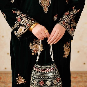 Pode incluir: Vestido de veludo verde escuro com intrincados bordados florais em dourado, vermelho e creme. O vestido apresenta mangas compridas com punhos detalhados e uma bolsa de mão a condizer. Pulseiras douradas e um anel completam o look.