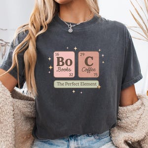Puede incluir: Camiseta gris oscuro con un diseño gráfico al estilo de la tabla periódica, con "Bo" y "C" que representan Libros y Café. El texto "The Perfect Element" está debajo. La camiseta se lleva con pantalones cortos vaqueros.