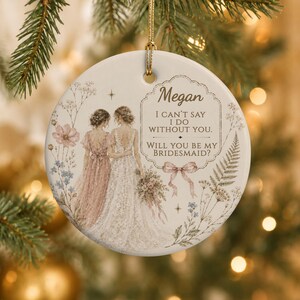 Puede incluir: Un adorno redondo de cer&aacute;mica blanca con una ilustraci&oacute;n de dos figuras con vestidos, uno rosa y otro blanco, rodeados de motivos florales. El texto dice: Megan, I CAN'T SAY I DO WITHOUT YOU. WILL YOU BE MY BRIDESMAID? con un lazo rosa.