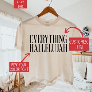 Könnte beinhalten: Ein cremefarbenes, kastenförmiges T-Shirt mit dem schwarzen Schriftzug "EVERYTHING HALLELUJAH" auf der Vorderseite. Das Bild enthält rote Textfelder mit den Phrasen "BOXY TEE", "CUSTOMIZE THIS!" und "PICK YOUR COLOR FONT" sowie rote Pfeile.