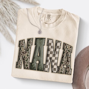 May include: Cream-colored Comfort Colors t-shirt with the word "NANA" in a collegiate style. The letters feature a camouflage pattern, a green hue, and a checkered design. A silver necklace with a pendant is draped on the shirt.
