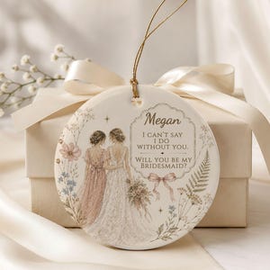 Puede incluir: Un adorno redondo de cer&aacute;mica blanca con una ilustraci&oacute;n de una novia y una dama de honor, rodeadas de motivos florales. El texto dice: Megan, I CAN'T SAY I DO WITHOUT YOU. WILL YOU BE MY BRIDESMAID? Cuelga de un cord&oacute;n dorado.