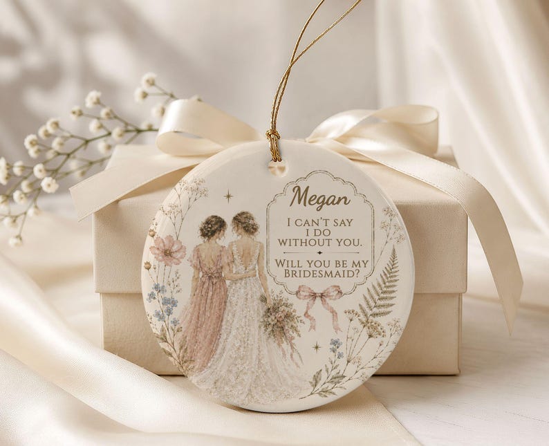 Puede incluir: Un adorno redondo de cer&aacute;mica blanca con una ilustraci&oacute;n de una novia y una dama de honor, rodeadas de motivos florales. El texto dice: Megan, I CAN'T SAY I DO WITHOUT YOU. WILL YOU BE MY BRIDESMAID? Cuelga de un cord&oacute;n dorado.