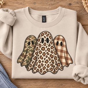 May include: A beige crewneck sweatshirt featuring a graphic of three ghosts. The left ghost has a green camouflage pattern, the center ghost has a brown leopard print, and the right ghost has a brown and white plaid pattern. Each ghost has two black oval eyes.