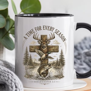 K&ouml;nnte beinhalten: Ein wei&szlig;er Keramikbecher mit schwarzem Henkel und Rand, verziert mit einer Grafik aus einem Holzkreuz, einem Hirsch, einer Ente und einem Fisch. Der Text A TIME FOR EVERY SEASON steht dar&uuml;ber und ECCLESIASTES 3:1 ist unter dem Motiv abgedruckt.
