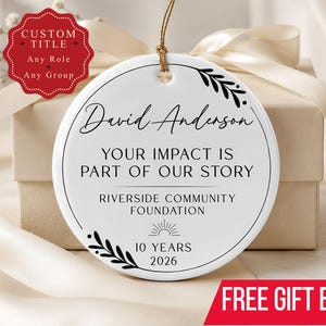 May include: A round white ceramic ornament featuring black text that reads David Anderson, YOUR IMPACT IS PART OF OUR STORY, RIVERSIDE COMMUNITY FOUNDATION, 10 YEARS, and 2026. The design includes black leaf accents and a sun icon, suspended by a gold cord.