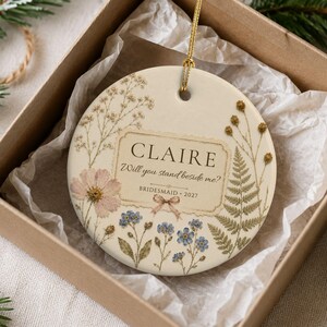 Puede incluir: Un adorno de cer&aacute;mica redondo de color crema con un cord&oacute;n dorado para colgar. La superficie muestra ilustraciones florales de estilo prensado, incluyendo flores rosas y azules y helechos verdes. El texto dice: CLAIRE, Will you stand beside me?, BRIDESMAID 2027.