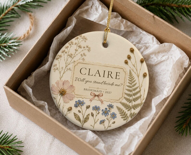 Puede incluir: Un adorno de cer&aacute;mica redondo de color crema con un cord&oacute;n dorado para colgar. La superficie muestra ilustraciones florales de estilo prensado, incluyendo flores rosas y azules y helechos verdes. El texto dice: CLAIRE, Will you stand beside me?, BRIDESMAID 2027.