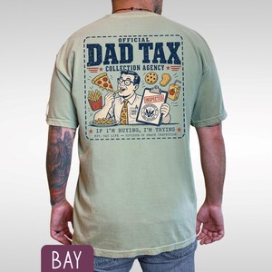 May include: Sage green t-shirt with a graphic on the back that reads "OFFICIAL DAD TAX COLLECTION AGENCY." The graphic features a cartoon man eating pizza and fries. The text "IF I'M BUYING, I'M TRYING" is also included.