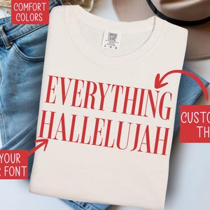 Könnte beinhalten: Ein cremefarbenes Kurzarm-T-Shirt mit dem roten Schriftzug EVERYTHING HALLELUJAH auf der Vorderseite. Das T-Shirt hat ein Markenetikett von Comfort Colors am Halsausschnitt. Rote Textfelder mit weißer Schrift weisen mit Anweisungen auf das Shirt.