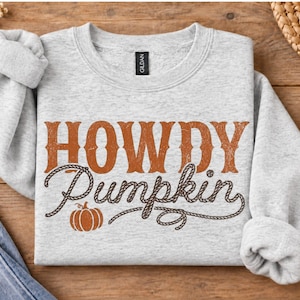 May include: A light gray crewneck sweatshirt featuring the orange distressed text HOWDY PUMPKIN across the chest. The word Pumpkin is written in a brown rope-style font, accompanied by a small brown pumpkin graphic at the bottom left corner.