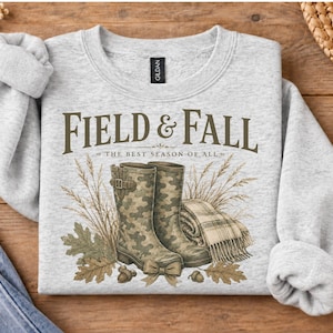 May include: A light gray crewneck sweatshirt featuring a graphic print of two camouflage rain boots, a plaid blanket, dried wheat stalks, and oak leaves. The text reads FIELD & FALL THE BEST SEASON OF ALL in dark brown lettering.