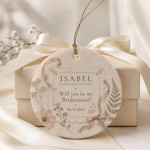 Puede incluir: Un adorno de cer&aacute;mica redondo de color crema con un lazo de hilo dorado. La superficie muestra ilustraciones florales, un gr&aacute;fico de cinta y el texto: ISABEL, For all the moments before the aisle, Will you be my Bridesmaid?, 06.12.2027.