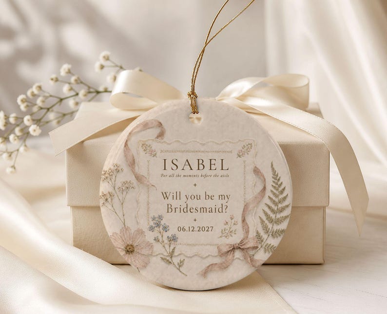 Puede incluir: Un adorno de cer&aacute;mica redondo de color crema con un lazo de hilo dorado. La superficie muestra ilustraciones florales, un gr&aacute;fico de cinta y el texto: ISABEL, For all the moments before the aisle, Will you be my Bridesmaid?, 06.12.2027.