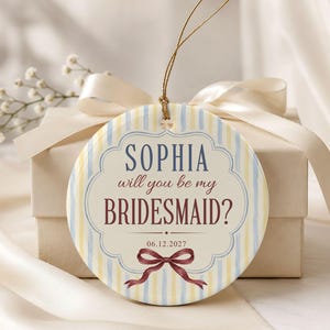 Puede incluir: Un adorno redondo de cer&aacute;mica con un patr&oacute;n de rayas verticales en azul claro y amarillo. El centro muestra el texto "SOPHIA will you be my BRIDESMAID?" en azul y granate, con la fecha "06.12.2027" y una ilustraci&oacute;n de un lazo granate.