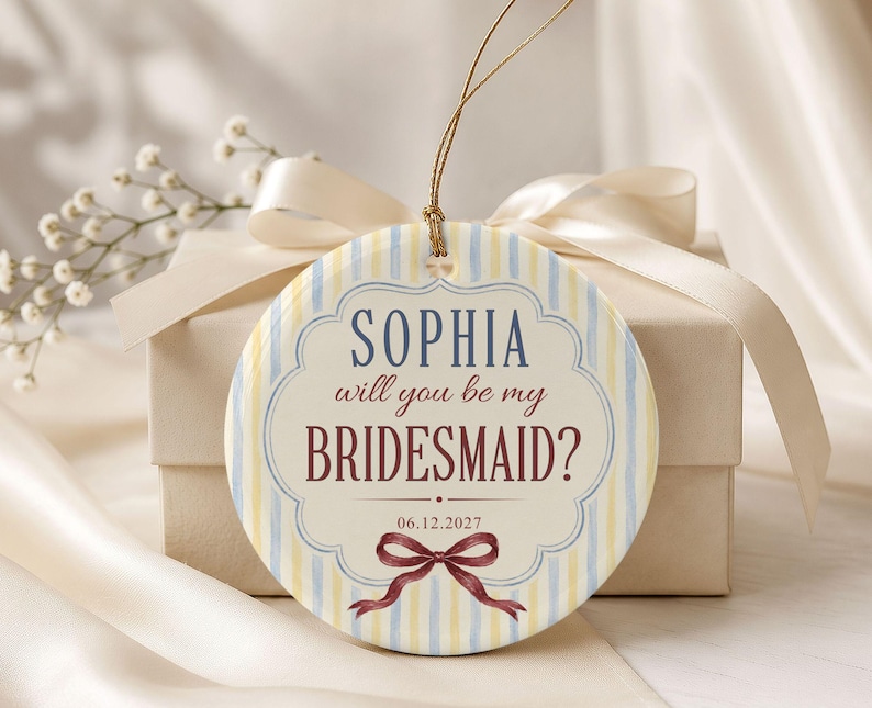 Puede incluir: Un adorno redondo de cer&aacute;mica con un patr&oacute;n de rayas verticales en azul claro y amarillo. El centro muestra el texto "SOPHIA will you be my BRIDESMAID?" en azul y granate, con la fecha "06.12.2027" y una ilustraci&oacute;n de un lazo granate.