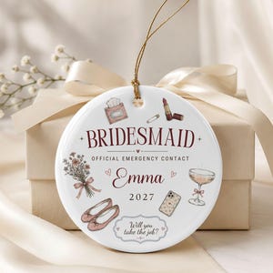 Puede incluir: Un adorno redondo de cer&aacute;mica blanca con el texto BRIDESMAID OFFICIAL EMERGENCY CONTACT, el nombre Emma, el a&ntilde;o 2027 y la frase Will you take the job? Las ilustraciones incluyen un ramo, pintalabios, pa&ntilde;uelos, bailarinas, un tel&eacute;fono y una copa.