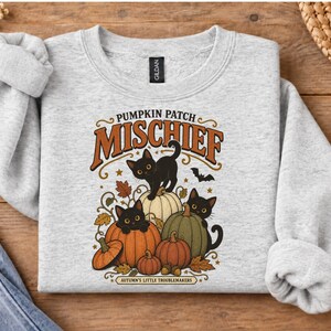 May include: A light gray crewneck sweatshirt featuring a graphic print of three black cats among pumpkins. The text reads PUMPKIN PATCH MISCHIEF and AUTUMN'S LITTLE TROUBLEMAKERS. The design includes orange, green, and white pumpkins with autumn leaves.