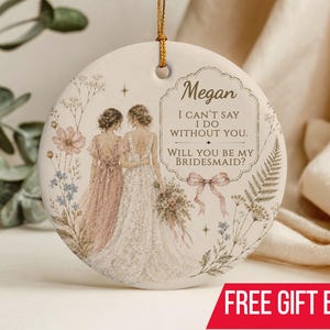 Puede incluir: Un adorno redondo de cer&aacute;mica blanca con un cord&oacute;n dorado para colgar. El dise&ntilde;o muestra dos figuras con vestidos, uno rosa y otro blanco, rodeadas de ilustraciones florales y el texto: Megan, I CAN'T SAY I DO WITHOUT YOU. WILL YOU BE MY BRIDESMAID?