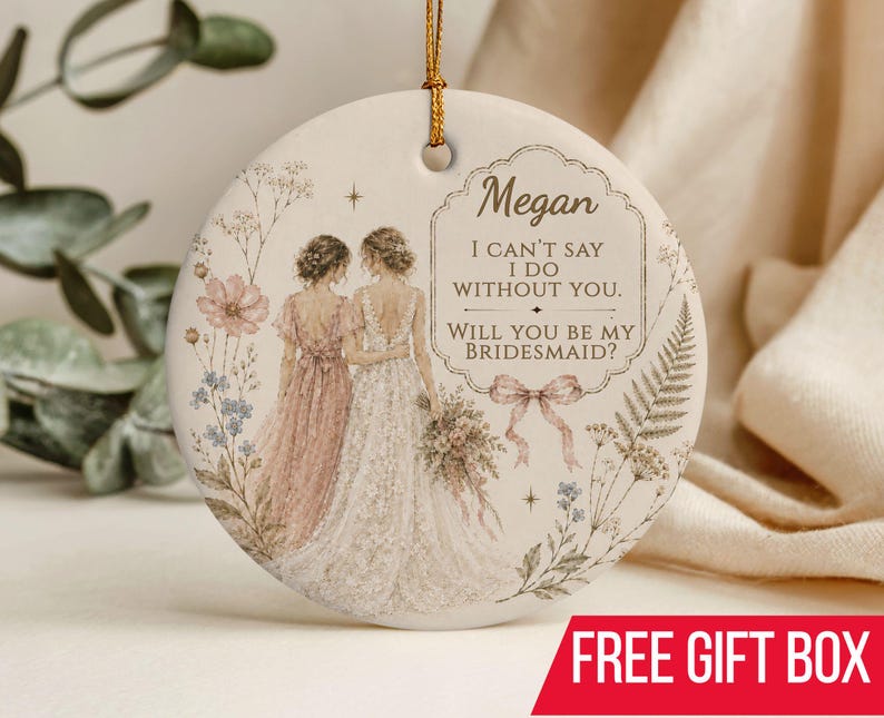Puede incluir: Un adorno redondo de cer&aacute;mica blanca con un cord&oacute;n dorado para colgar. El dise&ntilde;o muestra dos figuras con vestidos, uno rosa y otro blanco, rodeadas de ilustraciones florales y el texto: Megan, I CAN'T SAY I DO WITHOUT YOU. WILL YOU BE MY BRIDESMAID?