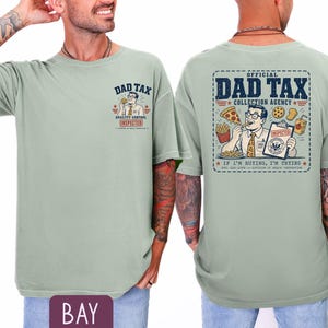 May include: A light green t-shirt with the text "DAD TAX" and a cartoon graphic on the front. The back of the shirt features a larger graphic with the words "OFFICIAL DAD TAX COLLECTION AGENCY" and the phrase "IF I'M BUYING, I'M TRYING."