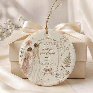 Puede incluir: Un adorno redondo de cer&aacute;mica blanca con un cord&oacute;n dorado, decorado con una ilustraci&oacute;n en acuarela de dos figuras con vestidos, motivos florales y el texto: CLAIRE Will you stand beside me? BRIDESMAID 2027, acentuado con un peque&ntilde;o lazo rosa.