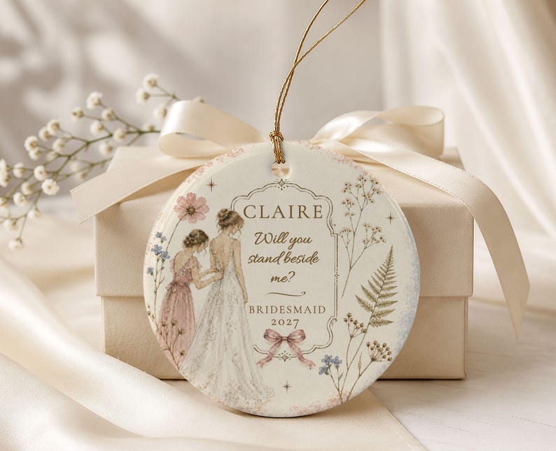 Puede incluir: Un adorno redondo de cer&aacute;mica blanca con un cord&oacute;n dorado, decorado con una ilustraci&oacute;n en acuarela de dos figuras con vestidos, motivos florales y el texto: CLAIRE Will you stand beside me? BRIDESMAID 2027, acentuado con un peque&ntilde;o lazo rosa.