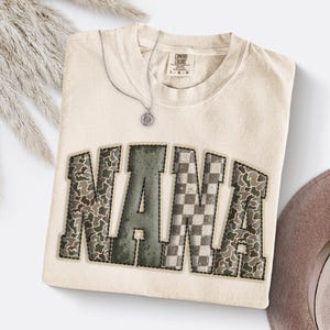 May include: Cream-colored Comfort Colors sweatshirt with the word "NANA" in a patchwork design. The letters feature a mix of camouflage and checkered patterns. A silver necklace with a pendant is draped on the shirt.
