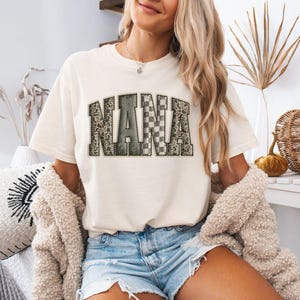 May include: Cream-colored t-shirt with the word "NANA" in a collegiate-style font. The letters have a mix of patterns, including a snakeskin print and a checkered design. The shirt is paired with denim shorts.