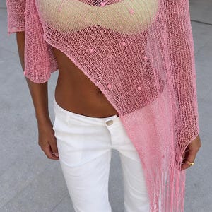 May include: A pink crochet top with a loose, open-knit texture and small pom-pom details, featuring an asymmetrical hem with fringe. The top is worn over a light-coloured bra and paired with solid white trousers, complemented by a gold-toned pendant necklace.