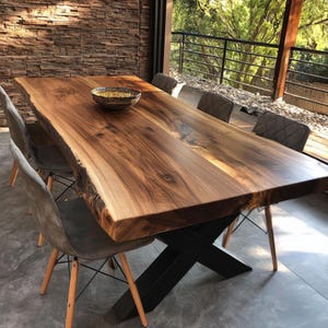 May include: A substantial wooden dining table with a live edge, supported by a black metal X-frame. Six grey upholstered chairs with wooden legs are arranged around the table. A decorative bowl sits on the table. The table is in a room with a stone wall and a view of trees.