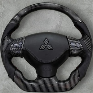 May include: A black and grey carbon fibre steering wheel with a Mitsubishi logo. The wheel has a perforated leather grip and integrated control buttons. The airbag is labelled "SRS AIRBAG".