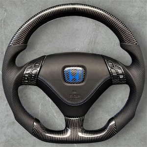 Pode incluir: Um volante preto e de fibra de carbono com um emblema azul da Honda no centro. O volante tem punhos de couro perfurado e botões de controle. O padrão de fibra de carbono é visível nos raios superior e inferior.