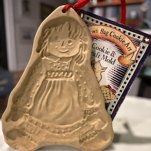 Könnte beinhalten: Eine hellbraune Keks- und Bastelform in Form eines Mädchens, das ein Kleid trägt. Die Form hat oben ein Loch zum Aufhängen und ist an einem Etikett mit dem Text "Brown Bag Cookie Art" befestigt. Ein rotes Band ist an der Form befestigt.