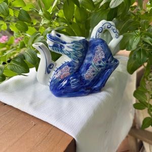 May include: A ceramic swan-shaped teapot with a blue and white floral design. The teapot features a curved handle and a spout, with blue accents on the wings and body. Pink floral details are painted on the body of the swan.