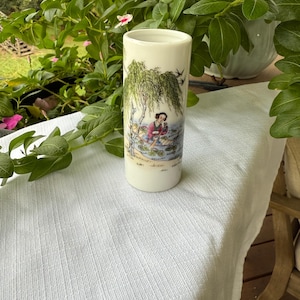 Könnte beinhalten: Zylindrische Keramikvase mit handgemalter asiatischer Szene. Das Design zeigt eine Frau in einem Boot unter einer Trauerweide. Die Vase ist cremefarben mit glatter Oberfläche und von Grün umgeben.