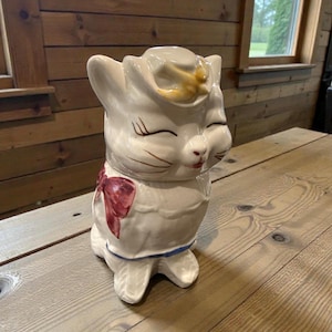 Pode incluir: Um pote de biscoitos de cerâmica branca em forma de gato com chapéu de cowboy e laço vermelho. O gato tem olhos fechados e bigodes castanhos. O pote tem uma borda azul e está em uma mesa de madeira.