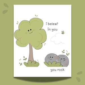 May include: A white greeting card featuring a light green tree with a smiling face, two grey rocks with smiling faces, a small bee, and a white cloud. The text reads I beleaf in you and you rock in a simple dark grey font on the white background.