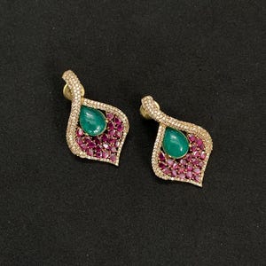 Peut inclure: Une paire de boucles d'oreilles en forme de goutte avec une pierre verte centrale entourée de petites pierres rose foncé et d'un bord de pierres blanches claires. Les boucles d'oreilles ont une monture en métal doré et une silhouette en forme de feuille.