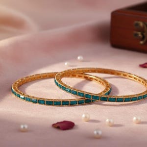 Puede incluir: Dos brazaletes dorados con piedras rectangulares de color verde azulado se exhiben sobre una superficie rosa. Los brazaletes están adornados con pequeñas piedras cuadradas verde azulado. Perlas y pétalos de rosa están dispersos.